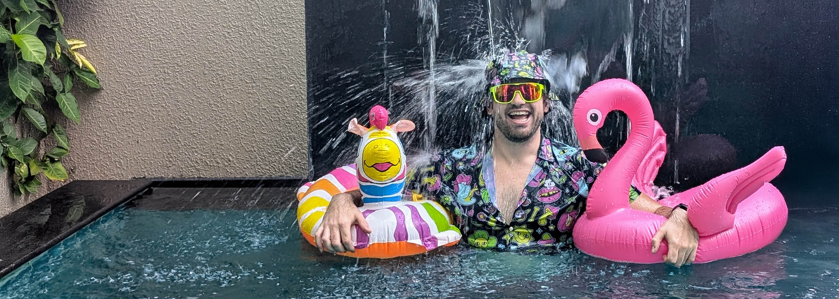 A man in colorful pool attire and sunglasses stands in a pool under a waterfall, smiling and holding inflatable floaties shaped like a pink flamingo and a rainbow unicorn. Green leaves hang on a nearby wall.