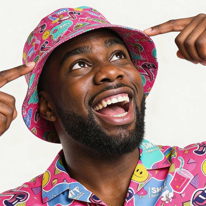 A smiling man points at his Badass Bucket Hat, which matches his shirt. Both feature playful designs—smiley faces, lightning bolts, and sunglasses—on a colorful pink background, perfect for festivals.