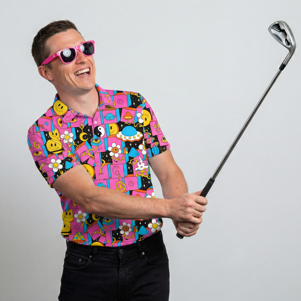 A smiling man in a Trippy Space Golf Polo Shirt and pink novelty sunglasses holds a golf club as if about to swing, standing against a plain light gray background.