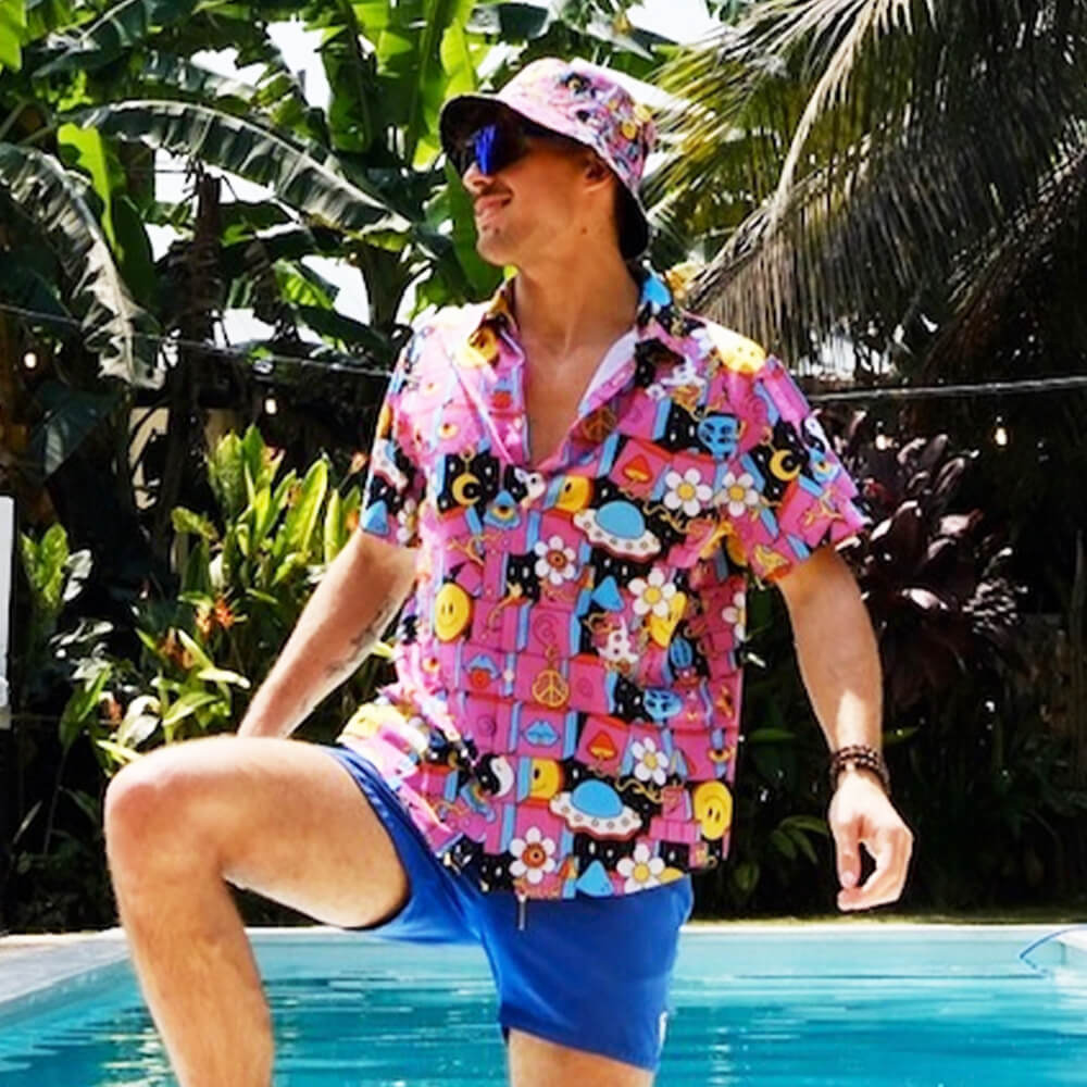 Wearing a Trippy Space Shirt, blue shorts, and a matching bucket hat, a person stands by the pool with one knee raised, surrounded by tropical plants and palm trees.
