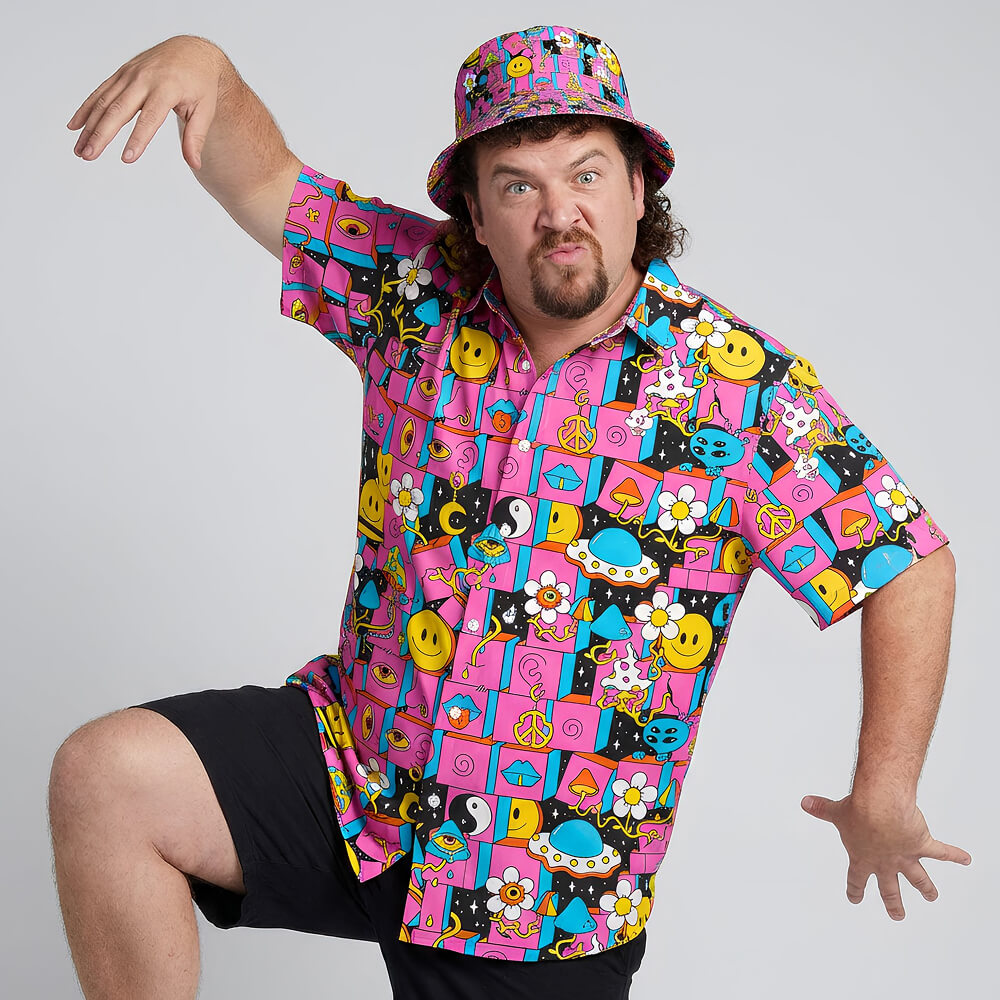A man in a magenta Trippy Space Shirt and matching bucket hat poses playfully with one arm raised and one leg bent. The shirt showcases smiley faces, flowers, peace signs, and quirky cartoon designs.