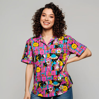 A smiling woman with curly brown hair stands against a plain gray background, wearing the Trippy Space Shirt—an eye-catching top with smiley faces, flowers, pandas, and geometric shapes.