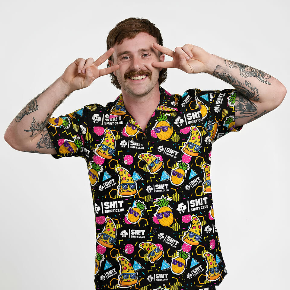 A tattooed man with a mustache smiles, flashing peace signs near his eyes. He wears the Wanna Pizza This Shirt, a colorful tee with burgers, fries, drinks, and fun text on a white background.