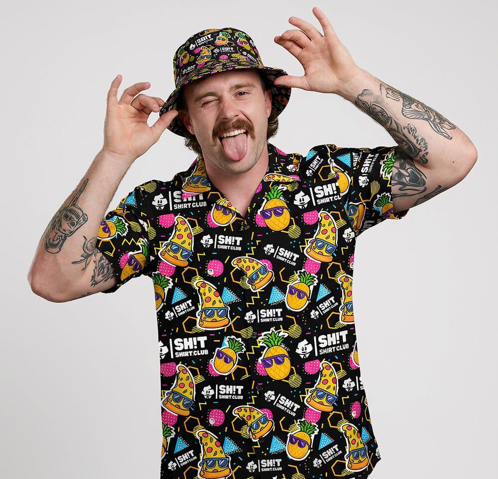 A tattooed man with a mustache and bucket hat sticks his tongue out while wearing the Wanna Pizza This Shirt, a colorful tee covered in cartoon food and drink graphics like burgers and sodas on a black background.