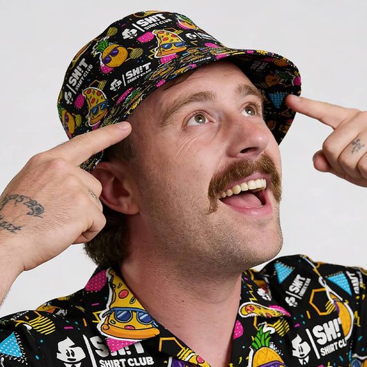 A smiling man with a mustache points to his Wanna Pizza This Bucket Hat, the perfect summer accessory. Both his shirt and hat feature vibrant cartoon graphics and “SHRT CLUB” text on a white background.