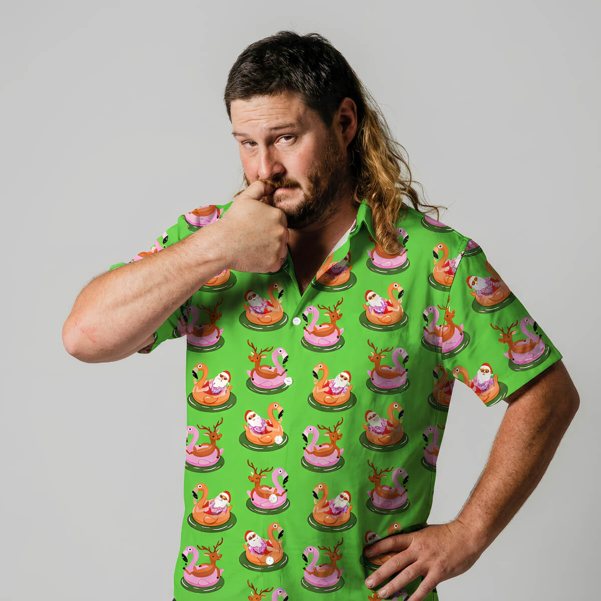 A man with brown hair and a beard stands against a gray background, looking thoughtful with his thumb on his chin. He’s wearing the bright green “Sleigh My Name, Sleigh My Name Shirt,” featuring cartoon flamingos in inflatable pool floats.