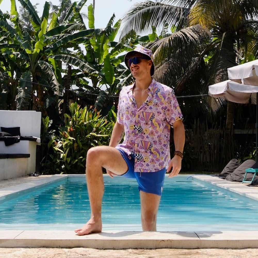 Wearing a Hippie Bucket Hat, a man in a colorful shirt and blue shorts smiles by the pool, with one knee on the edge, surrounded by lush tropical plants and palm trees.