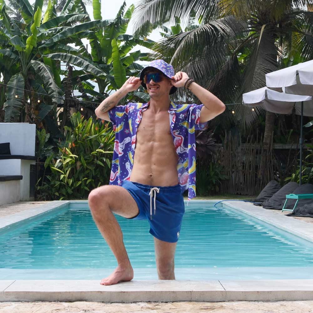 A man in a colorful shirt, blue shorts, and the Bananas For Flamingos Bucket Hat poses confidently by a swimming pool surrounded by tropical plants and palm trees.