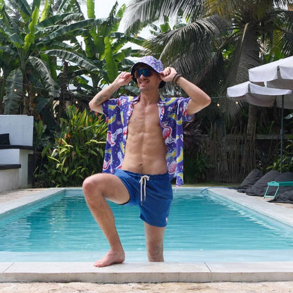 A man in a colorful shirt, blue shorts, and the Bananas For Flamingos Bucket Hat poses confidently by a swimming pool surrounded by tropical plants and palm trees.