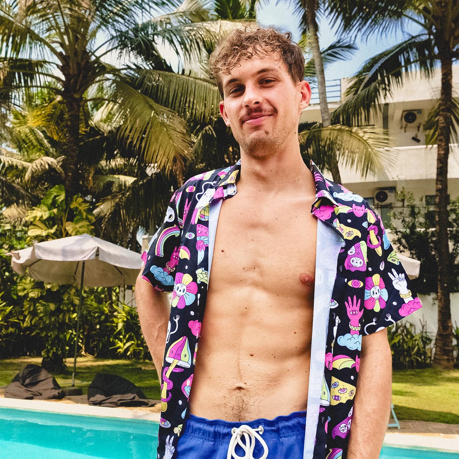A young man with curly hair stands poolside, wearing blue swim trunks and an open, colorful printed shirt, with palm trees and a building in the background on a sunny day.