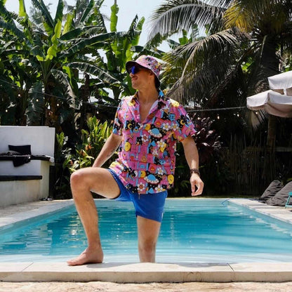 Wearing a colorful cartoon shirt, blue shorts, and the Trippy Space Bucket Hat, a man stands by a pool with one leg on the edge, surrounded by tropical plants and palm trees in bright sunlight—a perfect festival vibe.