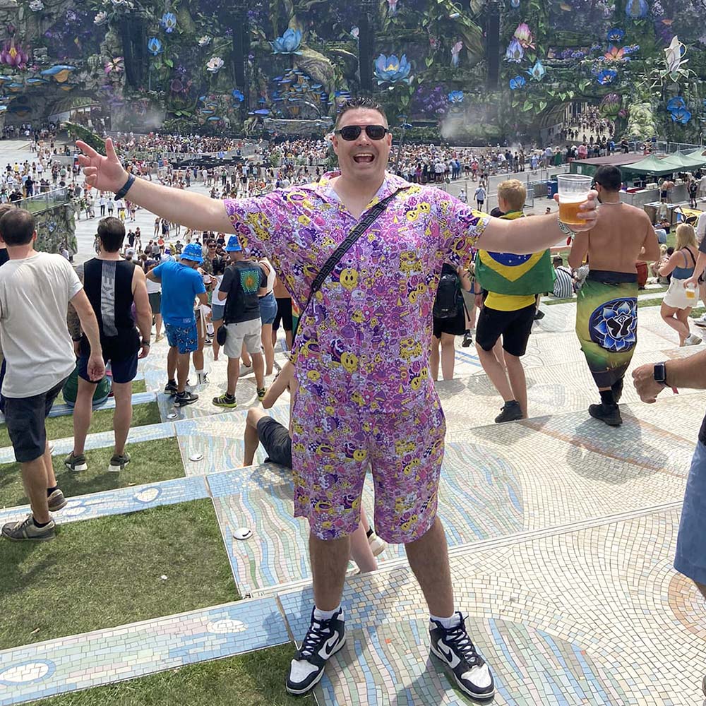 A man wearing a colorful, smiley-face patterned outfit stands with arms wide open, smiling at an outdoor music festival. He holds a drink, surrounded by crowds and vibrant stage decorations in the background.