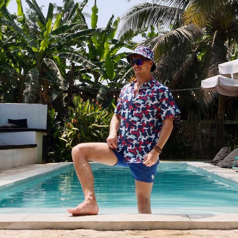 A man in a Combat2Coffee Army Camo Bucket Hat, sunglasses, a vibrant shirt, and blue shorts stands poolside with one leg raised, surrounded by lush tropical plants.