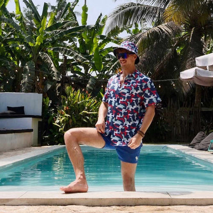 A man in a Combat2Coffee Army Camo Bucket Hat, sunglasses, a vibrant shirt, and blue shorts stands poolside with one leg raised, surrounded by lush tropical plants.