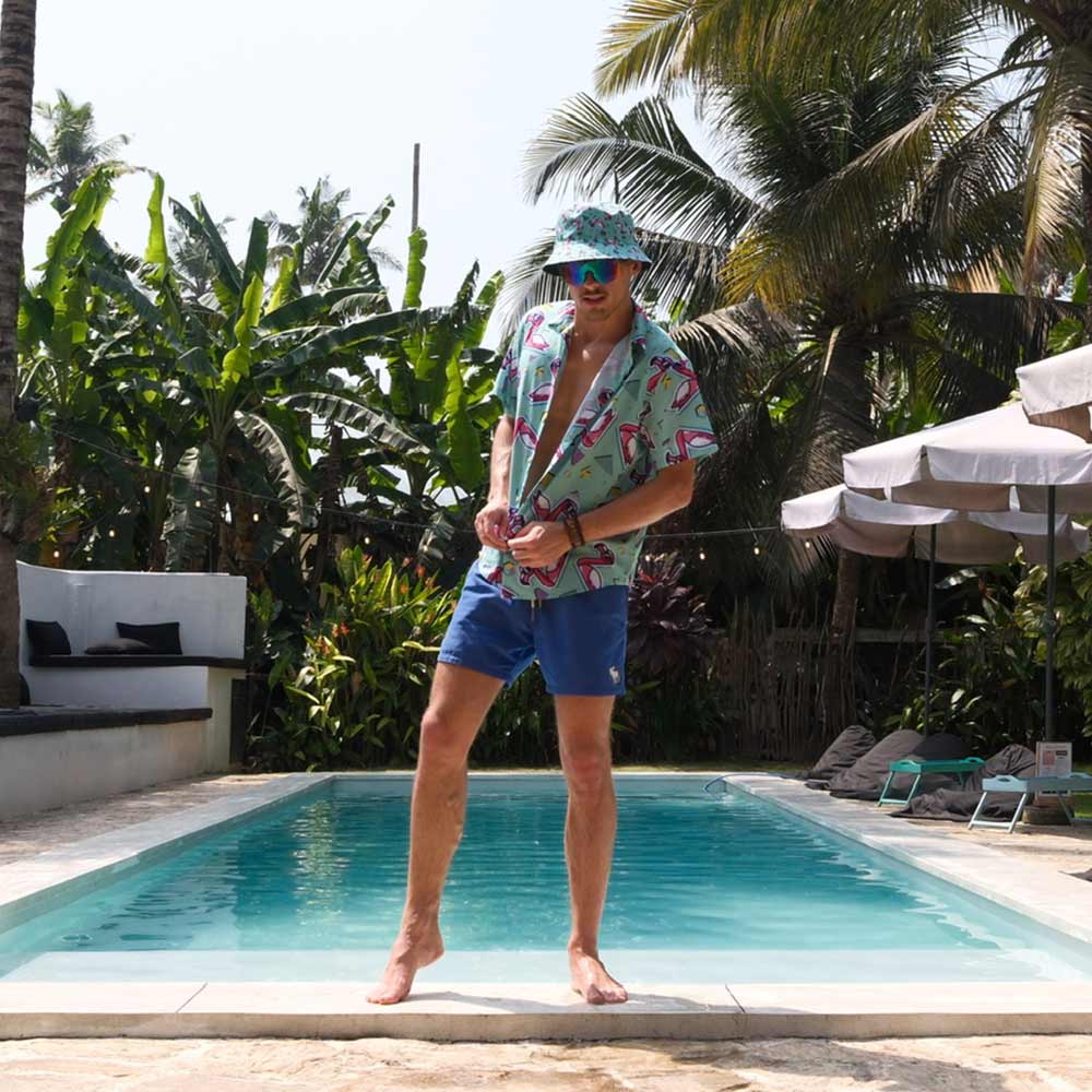 Wearing a Flamingo Fandango Bucket Hat, sunglasses, a flamingo-print shirt, and blue shorts, a man stands playfully at the edge of a tropical outdoor pool with palm trees and lounge chairs in the background.