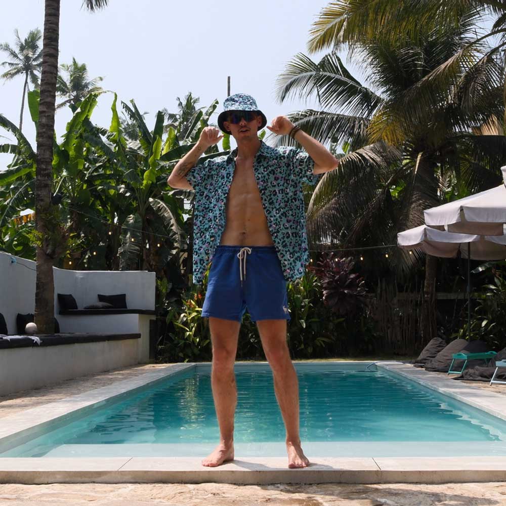 Wearing a Turquoise Tempest Leopard Print Bucket Hat, sunglasses, a patterned shirt, and blue swim trunks, a man stands confidently by the pool amid tropical plants and palm trees under the sunny sky.