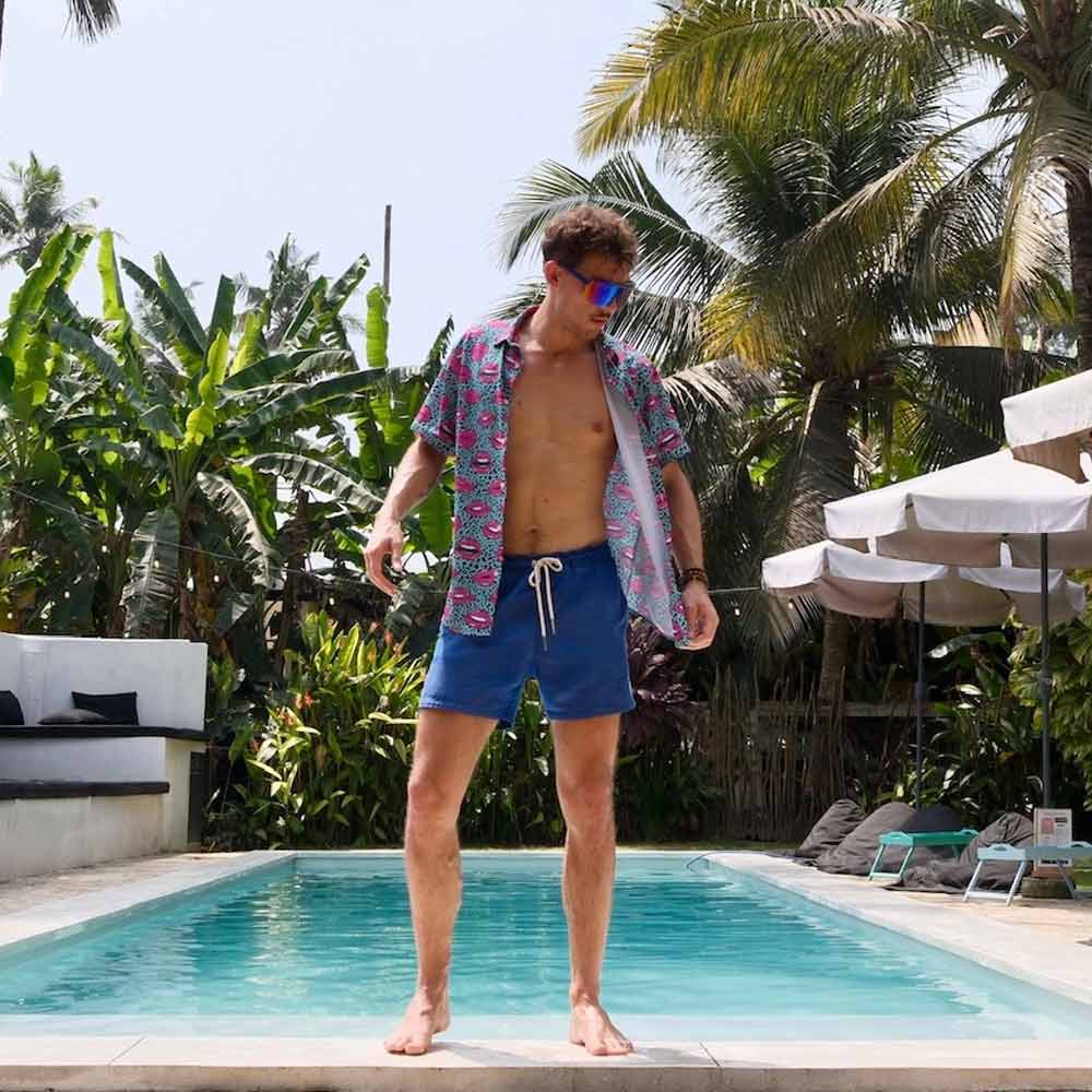 Wearing the Leopard Kiss Bad Bucket Hat, a man in blue swim trunks and a colorful open shirt stands poolside amid palm trees, lounge chairs, umbrellas, and lush greenery.
