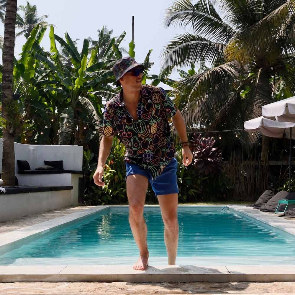 A man in a Fluorescent Fruit Fiesta Shirt, blue shorts, and a bucket hat steps out of a pool surrounded by tropical plants and palm trees, creating a relaxed, sunny vibe.
