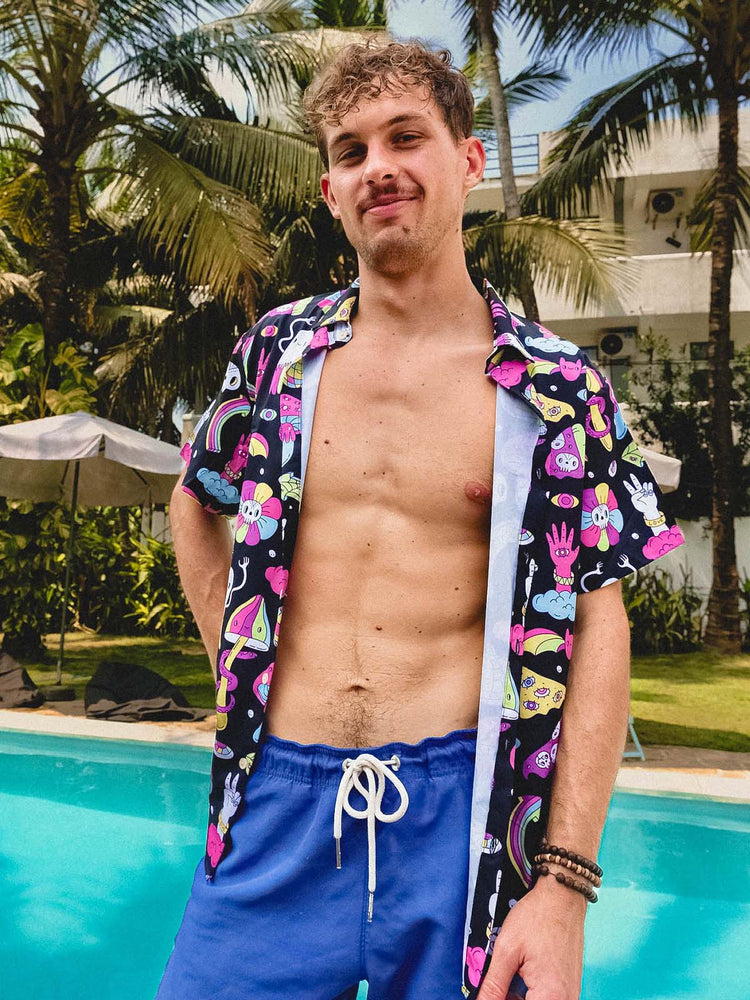 A young man with short curly hair stands by a pool, wearing blue swim trunks and an open, colorful shirt with cartoon prints. Palm trees and a white building are in the background. He is smiling and posing confidently.