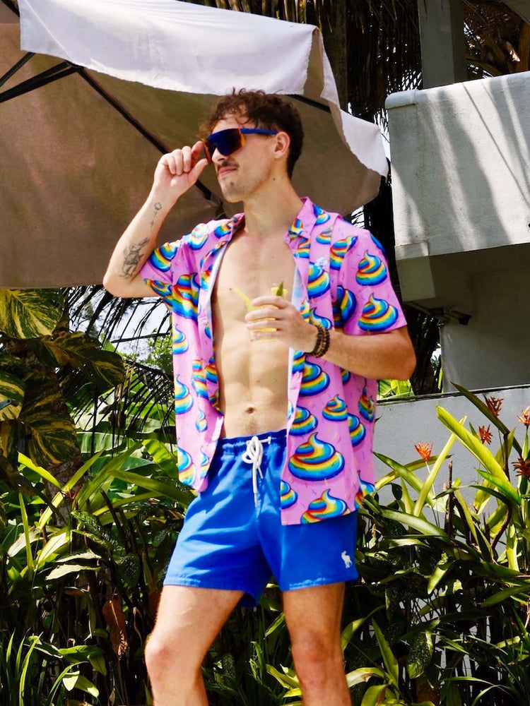 A man in sunglasses and a bright pink shirt with colorful swirl patterns stands outside by tropical plants, holding a drink and wearing blue swim trunks, with a large umbrella and building in the background.