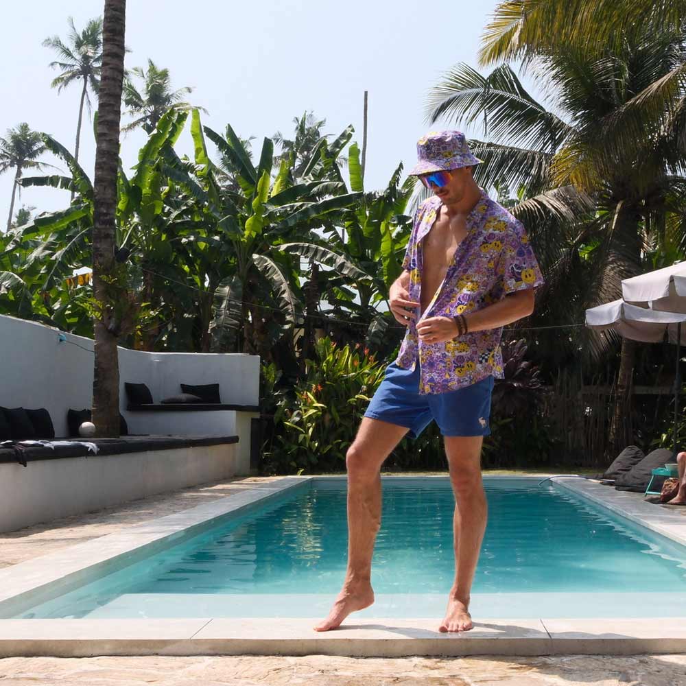 A man wearing the Magical Psychedelic Bucket Hat, a floral shirt, and blue shorts stands playfully by a sunny pool bordered by tropical plants and palm trees.