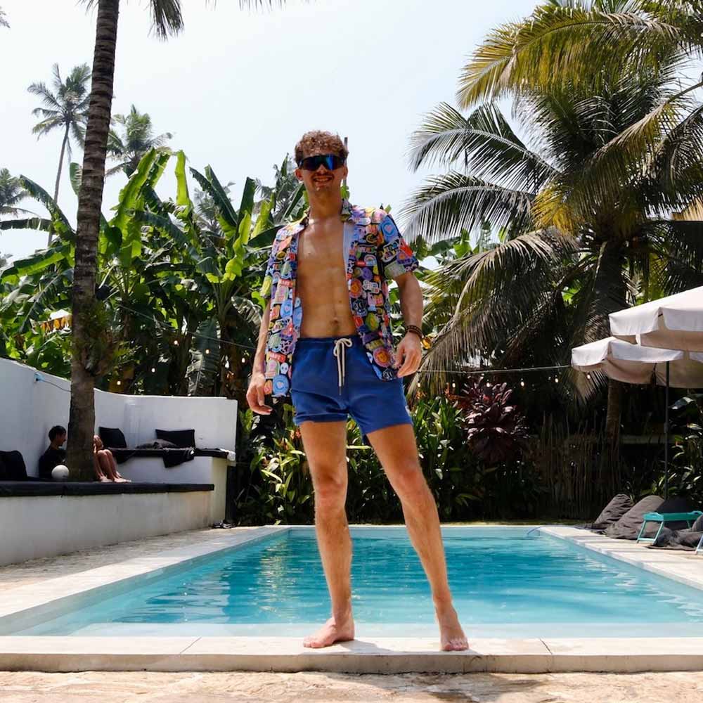 A man in blue swim trunks and a Retro Sticker Shirt stands by a pool, surrounded by lush tropical plants and palm trees, with people relaxing in the background.