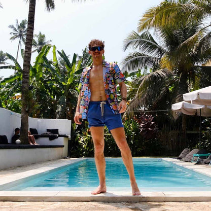 A man in blue swim trunks and a Retro Sticker Shirt stands by a pool, surrounded by lush tropical plants and palm trees, with people relaxing in the background.