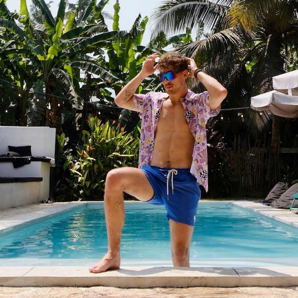 A man in blue swim shorts, a Hippie Shirt, and sunglasses stands by a sunny pool, posing with one knee up and adjusting his sunglasses. Lush tropical plants and palm trees surround the pool area.