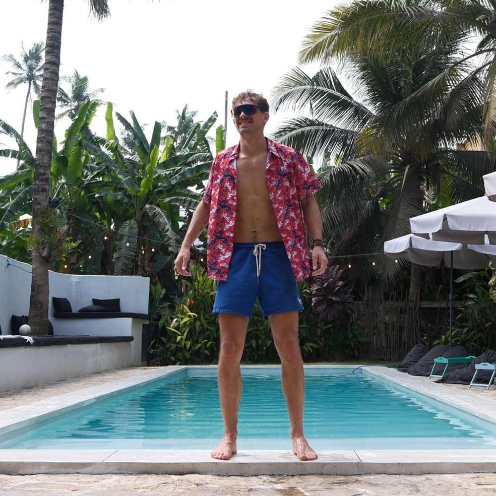 A man in a Red England FC Hawaiian Shirt and blue swim shorts stands by a pool amid tropical plants and palm trees, with outdoor lounge chairs and umbrellas in the background.