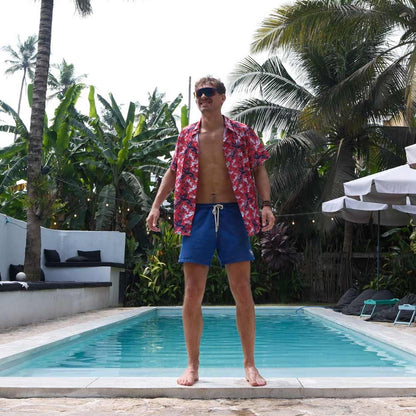 A man in a Red England FC Hawaiian Shirt and blue swim shorts stands by a pool amid tropical plants and palm trees, with outdoor lounge chairs and umbrellas in the background.