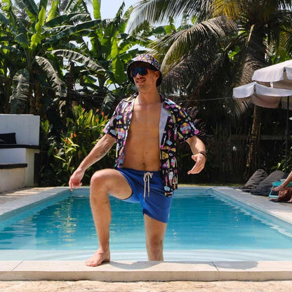 A man in a colorful shirt, blue shorts, sunglasses, and the Shroom Bucket Hat kneels at a pool’s edge with tropical plants and palm trees behind him.