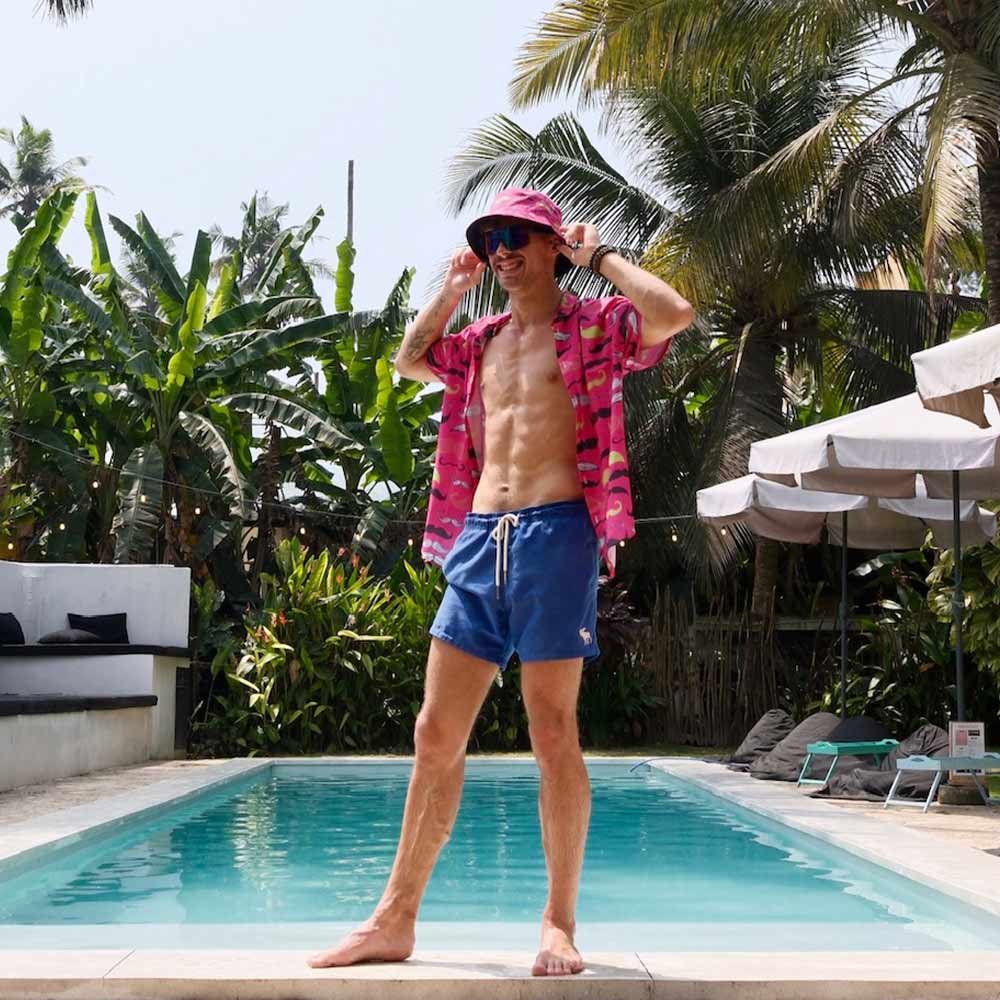 A man in blue swim shorts, a pink shirt, and a Movember Bucket Hat stands smiling by the pool, adjusting his hat. Palm trees and lush greenery surround the sunny pool area.