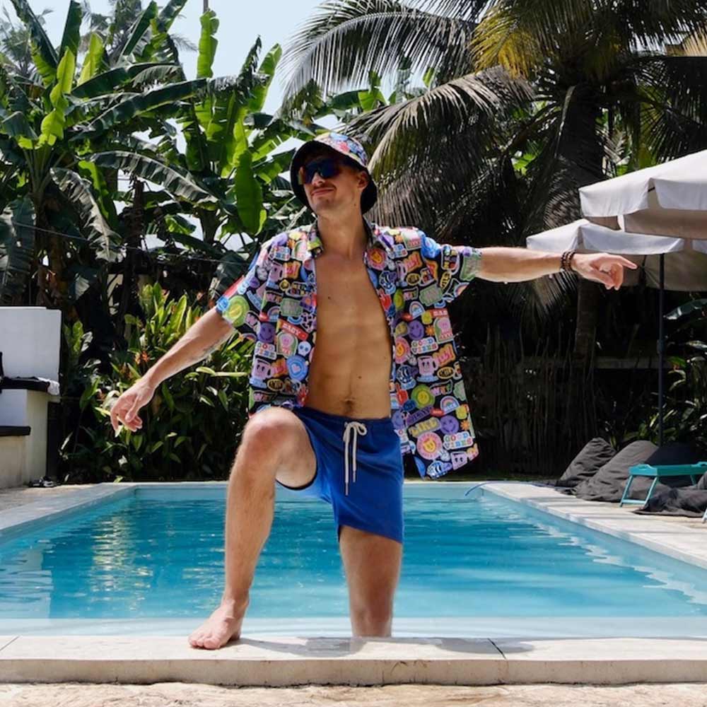 Wearing the Retro Sticker Bucket Hat, a man in a colorful shirt and blue shorts poses confidently with one knee up by a sunny pool, surrounded by lush tropical plants and palm trees.
