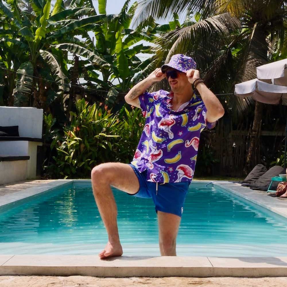 A man in vibrant swimwear and a Bananas For Flamingos Bucket Hat poses by a pool with one knee raised, surrounded by tropical plants and palm trees under a sunny sky.