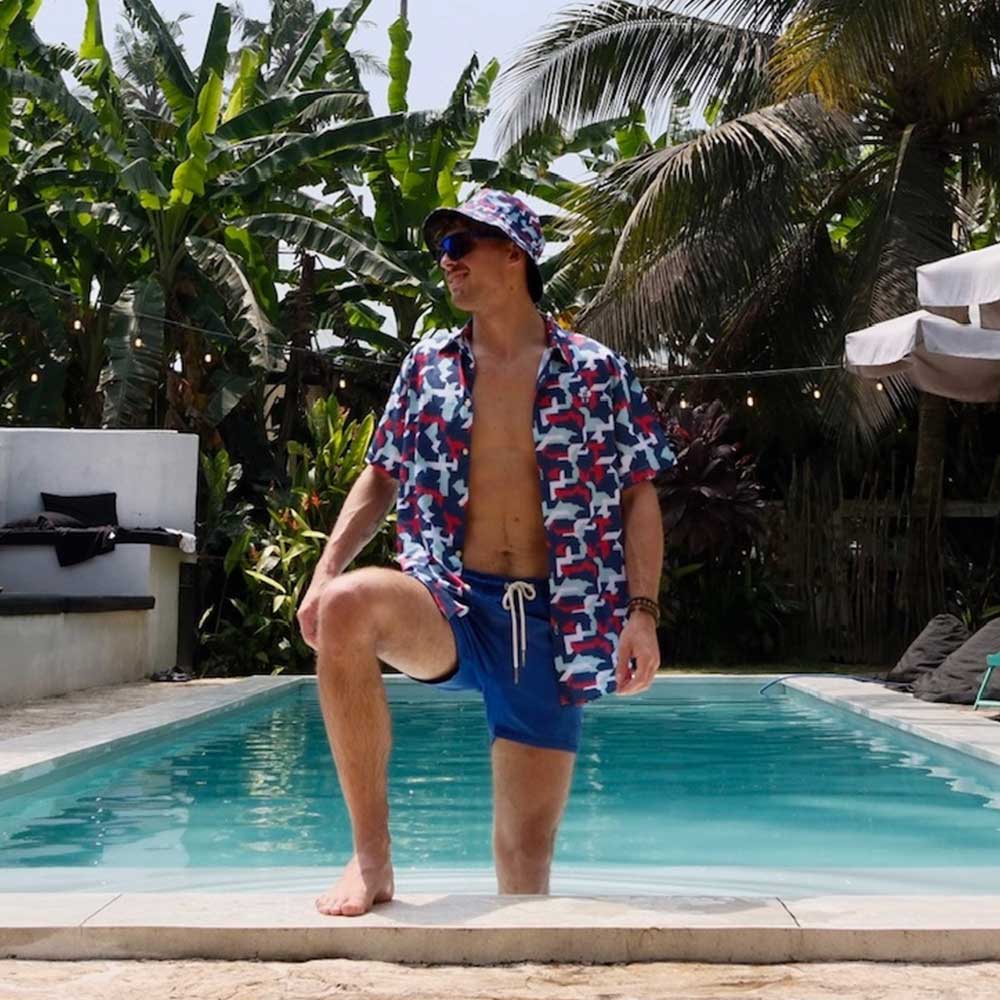 Wearing the Combat2Coffee Army Camo Bucket Hat and patterned swimwear, a person stands by the pool with one leg in the water, surrounded by tropical plants and palm trees on a sunny day.
