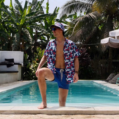 Wearing the Combat2Coffee Army Camo Bucket Hat and patterned swimwear, a person stands by the pool with one leg in the water, surrounded by tropical plants and palm trees on a sunny day.