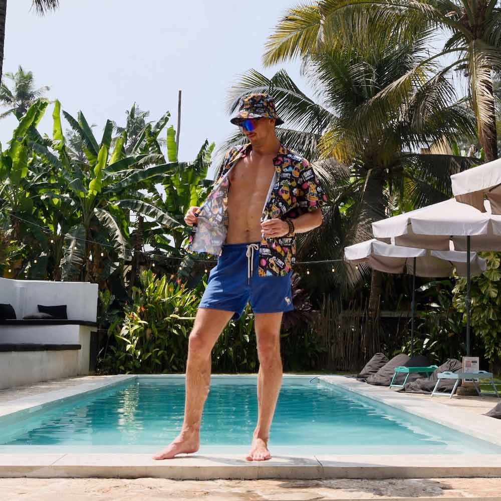 A man stands confidently at the edge of a pool, surrounded by tropical plants and lounge chairs, wearing a colorful Sh!t Shirt Club tee, blue swim shorts, and the Wanna Pizza This Bucket Hat.