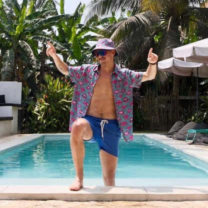 A man wearing sunglasses, blue shorts, an open patterned shirt, and the Leopard Kiss Bad Bucket Hat poses by a pool with one knee raised and both arms up. Palm trees and outdoor furniture are visible in the background.