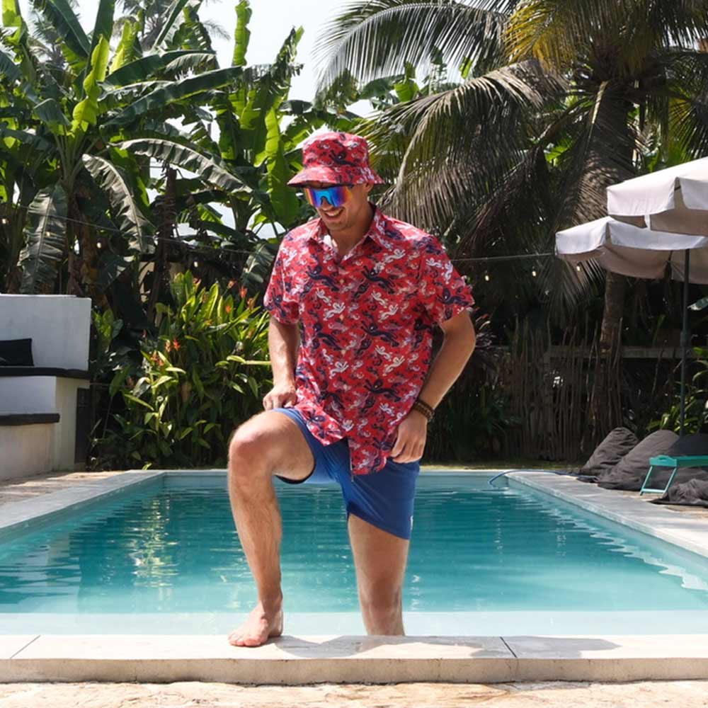 A man wearing a red floral shirt, blue shorts, sunglasses, and a Red England FC Away Bucket Hat steps out of a pool surrounded by tropical plants and palm trees.