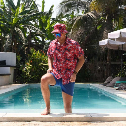 A man wearing a red floral shirt, blue shorts, sunglasses, and a Red England FC Away Bucket Hat steps out of a pool surrounded by tropical plants and palm trees.