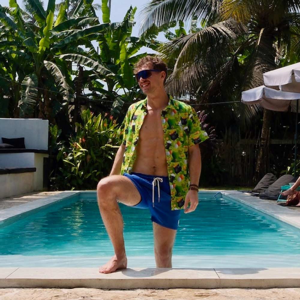 A man in a St Patricks Day Shirt, sunglasses, and blue swim shorts stands smiling by the pool. Palm trees and lush greenery frame the sunny scene.