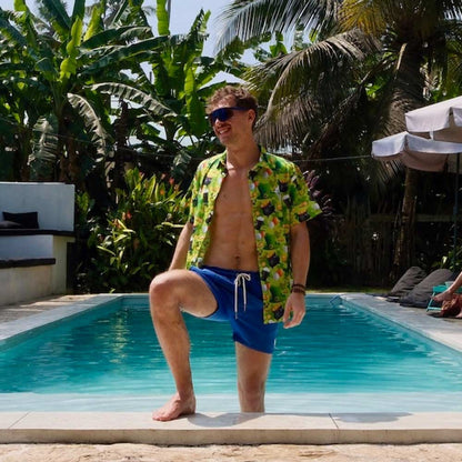 A man in a St Patricks Day Shirt, sunglasses, and blue swim shorts stands smiling by the pool. Palm trees and lush greenery frame the sunny scene.