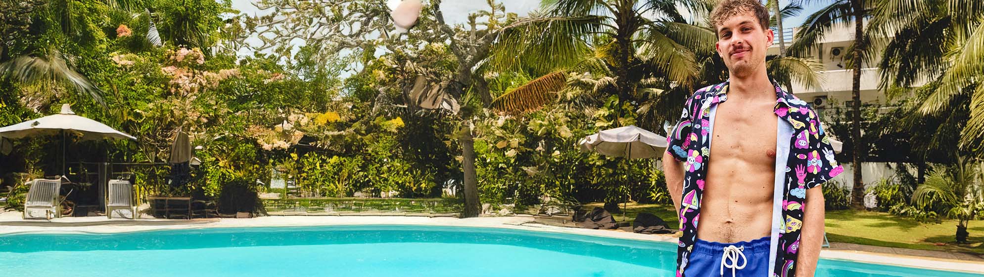 A young man wearing a patterned open shirt and blue swim trunks stands by a bright blue outdoor pool, surrounded by palm trees and greenery on a sunny day.
