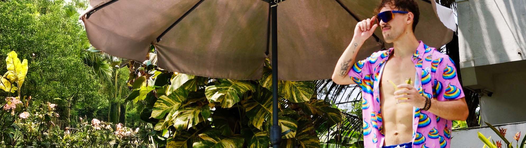 A man wearing a colorful shirt, swim trunks, and sunglasses stands under a large outdoor umbrella, holding a drink. He is outside near lush tropical plants and a building, enjoying a sunny day.