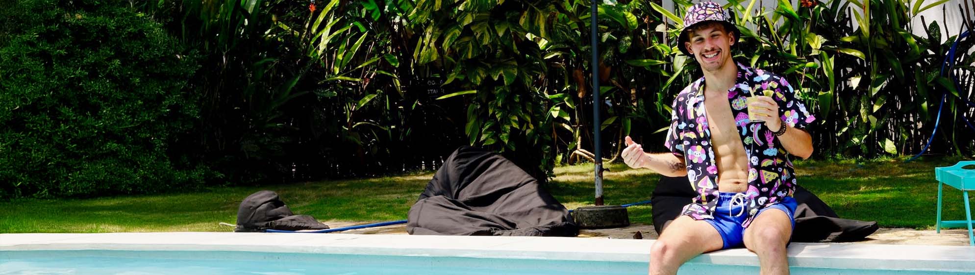 A smiling man sits poolside wearing a colorful shirt, matching bucket hat, and blue swim trunks, holding a drink and a smartphone. Lush greenery fills the background.
