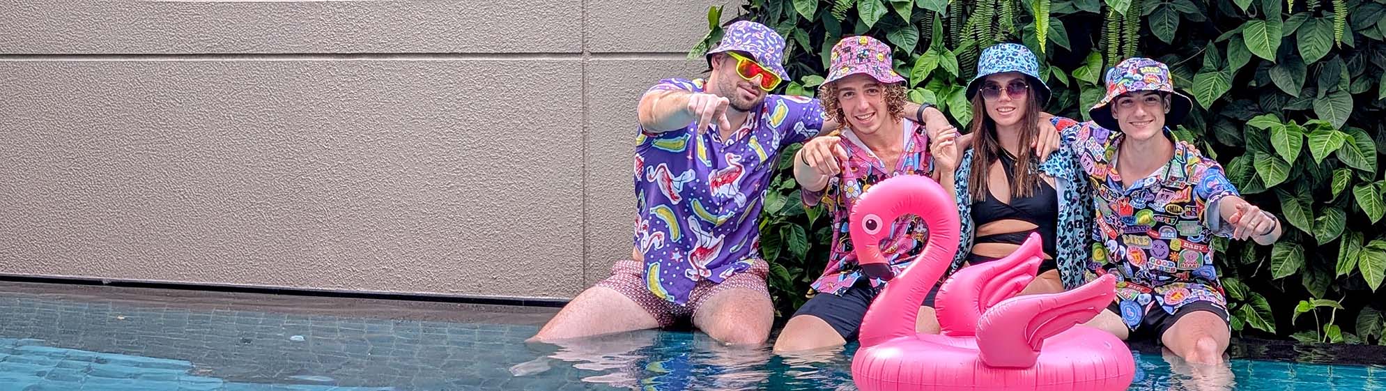 Four people in colorful matching outfits and bucket hats pose by a pool with a large pink inflatable flamingo. Green foliage and a beige wall are in the background.