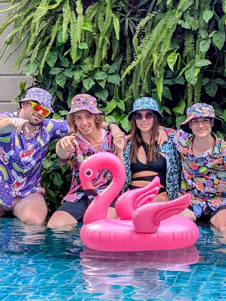 Four young adults in colorful, patterned outfits and bucket hats sit at the edge of a pool with a pink inflatable flamingo. Lush green plants are in the background, and everyone is smiling and having fun.