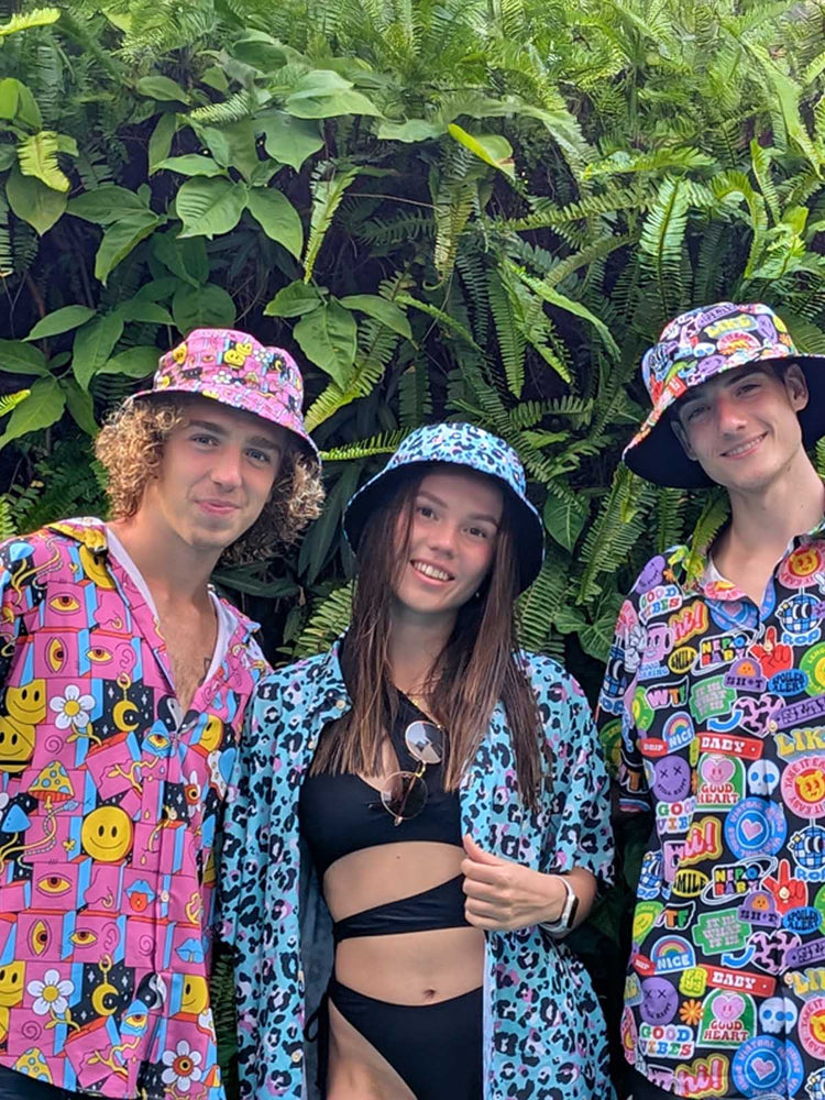 Three young adults pose in front of lush green ferns. Two men wear colorful bucket hats and patterned shirts, while the woman in the center wears a black swimsuit with a blue animal print shirt and a matching bucket hat. All are smiling.