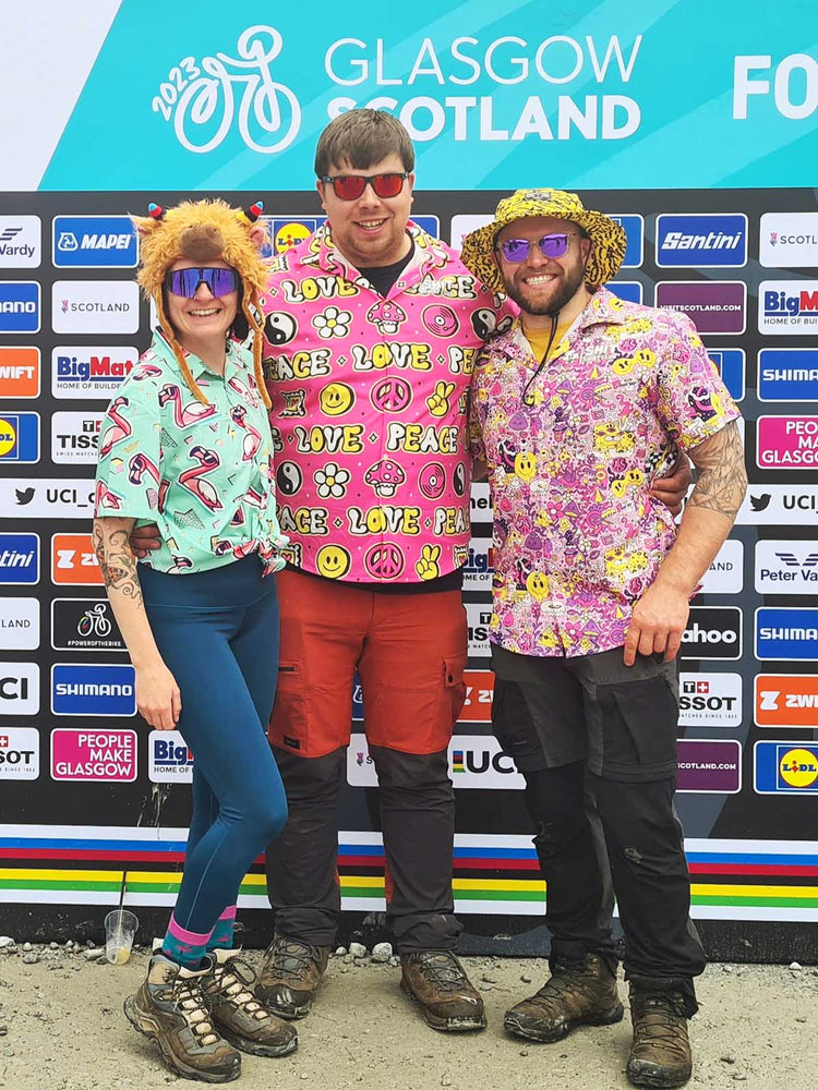 Three people in colorful, playful shirts and hats stand smiling in front of a backdrop with sponsor logos at the 2023 UCI Cycling World Championships in Glasgow, Scotland.