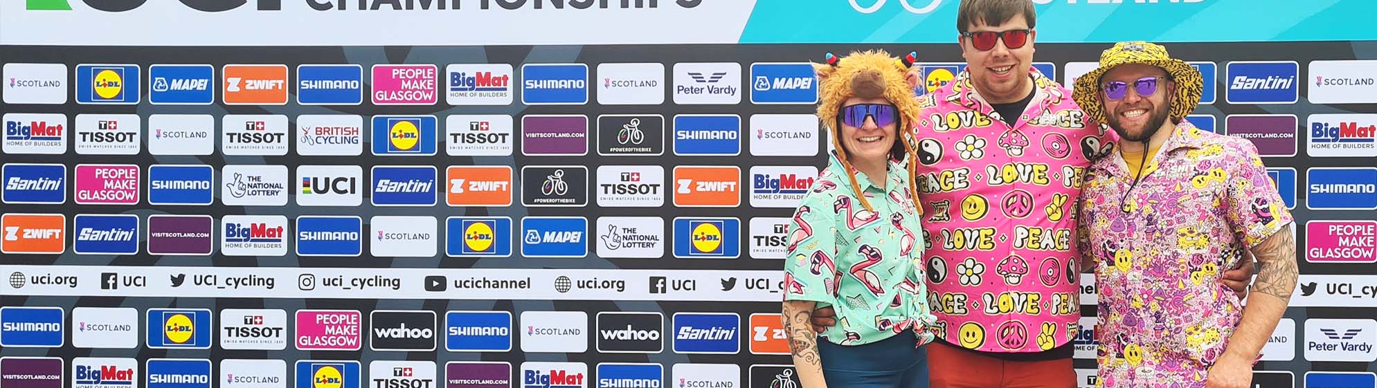 Three people in colorful, playful outfits and sunglasses stand smiling in front of a sponsor board at a cycling championship event. The background features multiple sponsor logos and event branding.